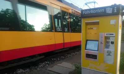 Das Foto zeigt einen gelb-weißen Fahrscheinautomaten der neuen Generation an einer AVG-Haltestelle. Im Hintergrund fährt eine Stadtbahn vorbei.