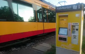 Das Foto zeigt einen gelb-weißen Fahrscheinautomaten der neuen Generation an einer AVG-Haltestelle. Im Hintergrund fährt eine Stadtbahn vorbei.