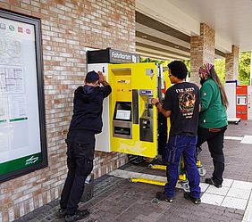 Drei Männer hieven einen neuen gelb-weißen Fahrscheinautomaten in eine Aussparung in einer Mauer am Bahnsteig der Haltestelle Böckingen Sonnenbrunnen