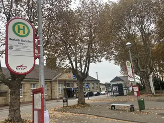 Das Foto zeigt den Vorplatz des Durlacher Bahnhofes. Im Vordergrund ist ein Haltestellenschild zu sehen, im Hintergrund Bäume und das Bahnhofsgebäude.
