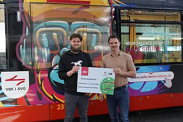 Das Foto zeigt Stefan Mock, Ausbildungsleiter bei VBK und AVG, bei der symbolischen Übergabe eines Deutschlandtickets an Michael aus Bad Herrenalb, der an der Fahrt mit Karaokebahn im April teilgenommen hatte. Beide stehen vor der Karaokebahn im Betriebshof der Verkehrsbetriebe.