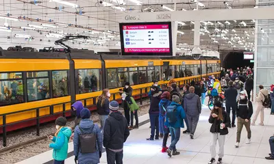 Fahrgäste stehen an einem Bahnsteig im Karlsruher Stadtbahntunnel. In der Bildmitte ist ein Haltestellen-Anzeiger für die fahrgastinformation zu sehen. Im Hintergrund fährt eine Bahn in die Haltestelle ein.