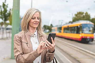 Eine Frau steht in einer Haltestelle und hält ein Smartphone in der Hand. Im Hintergrund ist eine Bahn zu sehen.