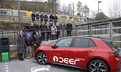 Das Foto zeigt ein rotes Elektroauto des Charsharinganbieters deer vor dem barrierfreien Zugang zur neu gestalteten Stadtbahnhaltestelle. Hinter dem Auto stehen Kämpfelbachs Bürgermeister Thomas Maag (2.von rechts.) und die Projektpartner, Vertreter*innen des Gemeinderats und Bürger*innen der Gemeinde.  