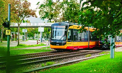 Eine Bahn der AVG-Linie S11 fährt durch den Karlsruher Stadtteil Rüppurr. Der Ast eines Laubbaumes ragt im Vordergrund in das Bild.