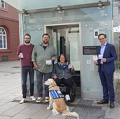 Vor dem Stadtbahn-Tunnel am Marktplatz sind von links nach rechts folgende Personen zu sehen: Christian Höglmeier (technischer Geschäftsführer der VBK), Ulrike Wernert (kommunale Behindertenbeauftragte der Stadt Karlsruhe), Artur Bundnik (Projektstelle Web-App „Karlsruhe barrierefrei“) und Marcus Albert (System-Administrator „Karlsruhe barrierefrei“). Sie halten die Aufkleber mit den QUR-Codes zu "Karlsruhe barrierefrei" in der Hand. Vor ihnen sitzt ein Hund. 