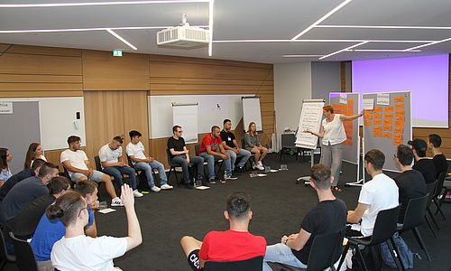 Junge Menschen sitzen in einem Stuhlkreis in einem Seminarraum und diskutieren mit einer Referentin über das Thema Nahchhaltigkeit.