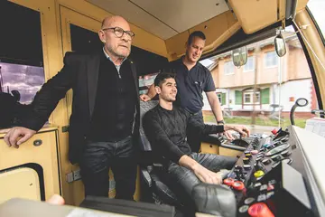 Landes-Verkehrsminister Herrmann begleitet im Führerstand einer AVG-Stadtbahn eien Schulungsfahrt. Auf dem Fahrersitz beindet sich ein Teilnehmer des Kurses "Qualifizierung Geflüchteter zu Triebfahrzeugführern". Hinter dem Kursteilnehmer steht ein Ausbilder der AVG.