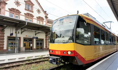 Eine AVG-Stadtbahn hält an einem Bahnsteig. Im Hintergrund ist ein Bahnhofsgebäude zu sehen.