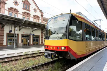 Eine AVG-Stadtbahn hält an einem Bahnsteig. Im Hintergrund ist ein Bahnhofsgebäude zu sehen.