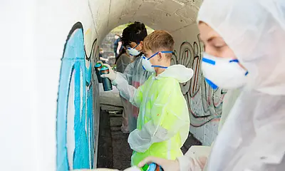 Schüler*innen gestalten mit Spraydosen die Unterführung an der Bahnhaltestelle in Baiersbronn. Dabei tragen die Schüler*innen Schutzkleidung.