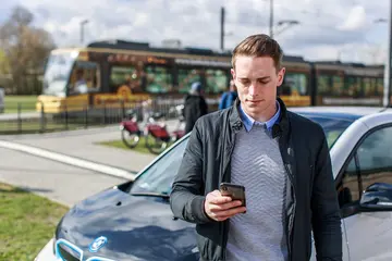 Ein junger Mann steht mit einem Smartphone in der Hand vor einem Carsharing-Auto. Im Hintergrund fährt eine Straßenbahn vorbei.