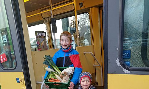 Zwei Kinder mit einem Obst- und Gemüsekorb in der Hand schauen aus einer geöffneten tür einer AVG-Stadtbahn