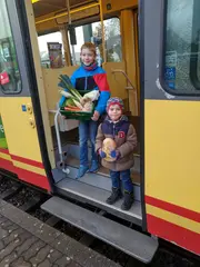 Zwei Kinder mit einem Obst- und Gemüsekorb in der Hand schauen aus einer geöffneten tür einer AVG-Stadtbahn