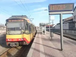 Eine gelb-rote Stadtbahn der AVG steht auf einem Gleis an der Haltestelle Freudenstadt Stadt. Rechts von der Bahn befindet sich der Bahnsteig mit einem Fahrgaszanzeiger, Mülleimer und Wartehäuschen. Der Himmel ist hellblau und leicht bewölkt.