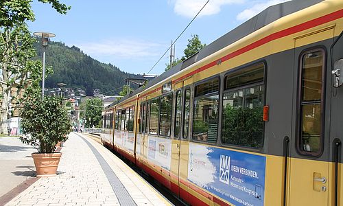 Rechts im Bild befindet sich eine AVG-Stadtbahn an einer Haltestelle in Bad Wildbad. Links ist der Bahnsteig zu sehen. Im Hintergrund befinden sich grüne, bewaldete Hügel des Schwarzwaldes.