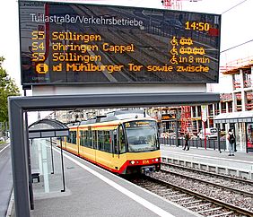 Eine digitale Fahrgastanzeige zeigt verschiedene Bahnen und deren Abfahrtszeiten. Im Hintergrund ist eine Stadtbahn an einer Haltestelle zu sehen.