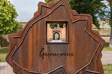 Rostbrauner, künstlerischer Aufsteller mit der Aufschrift "Germersheim". Im Hintergrund ist die Germersheimer Festung zu erkennen.