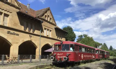 Fas Foto zeigt den roten Schienenbus, der in den Sommermonaten nach Maulbronn fährt. Im Hintergrund ist ein Bahnhofsgebäude zu sehen. 