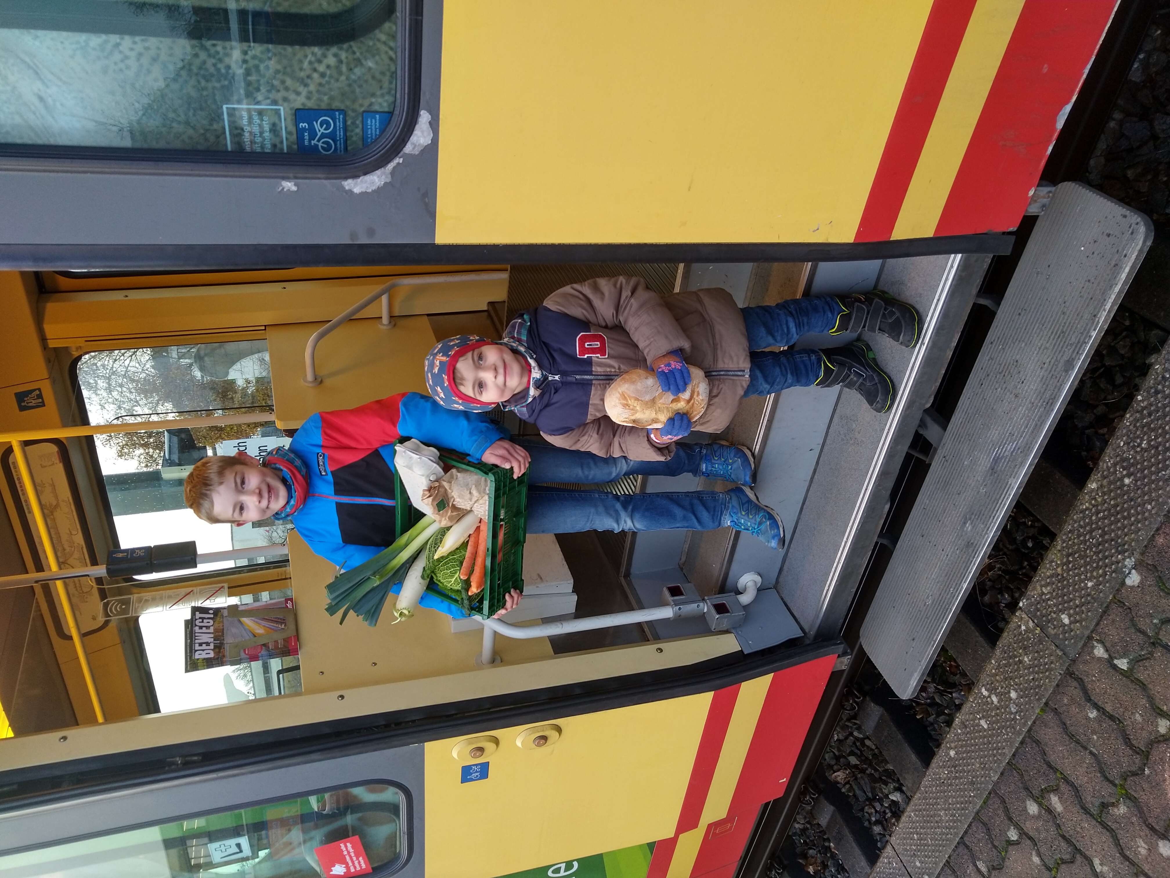 Zwei Kinder mit einem Obst- und Gemüsekorb in der Hand schauen aus einer geöffneten tür einer AVG-Stadtbahn